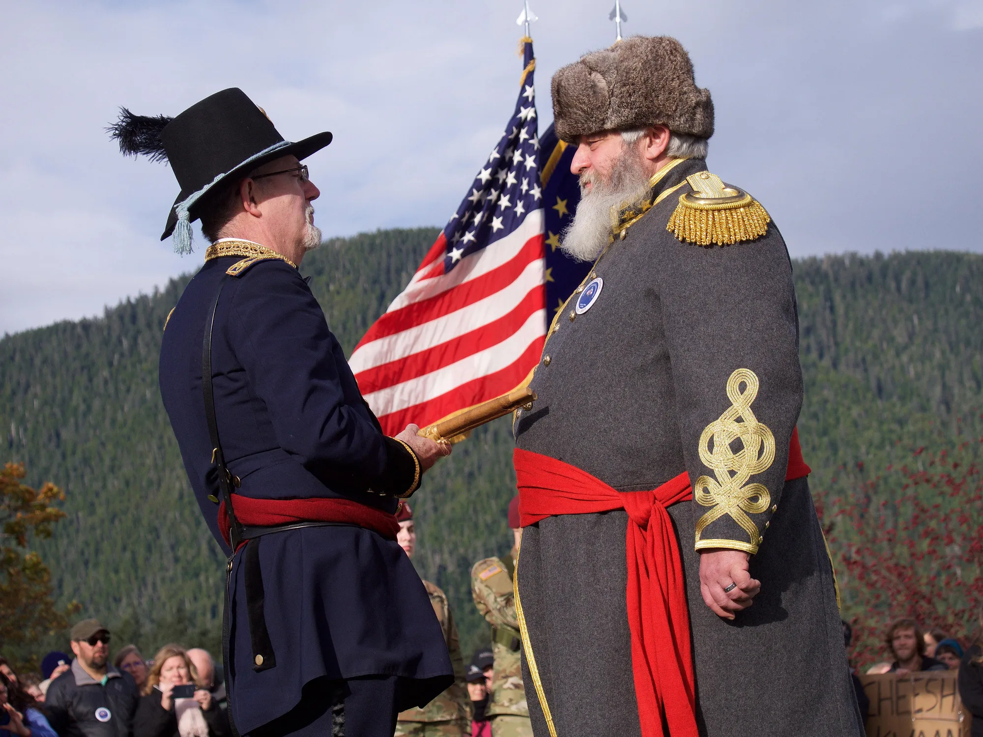 Two men in 19th-century uniforms face each other in front of an American flag, one in a plumed black hat and blue coat, the other with a white beard, fur hat and grey greatcoat with gold epaulettes, with a forested mountain behind.