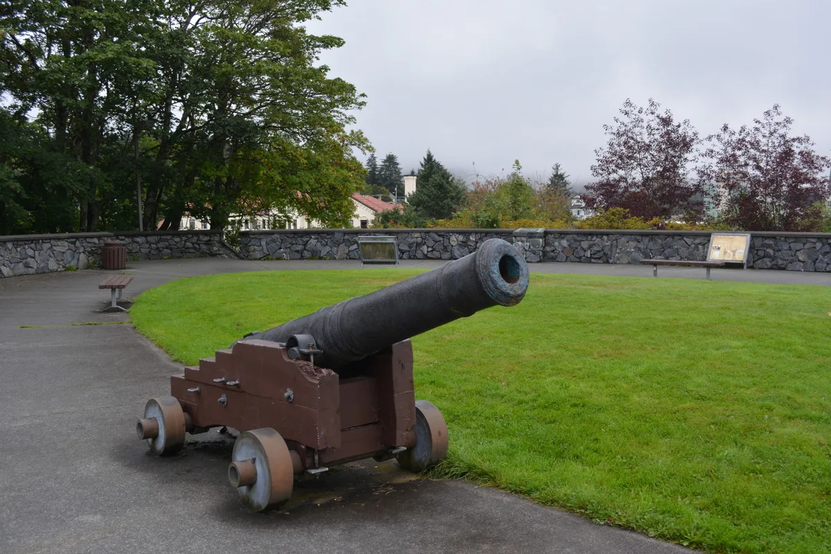 An iron cannon on a wooden wheeled carriage stands on paving beside a lawn, with a low stone wall and trees behind it.