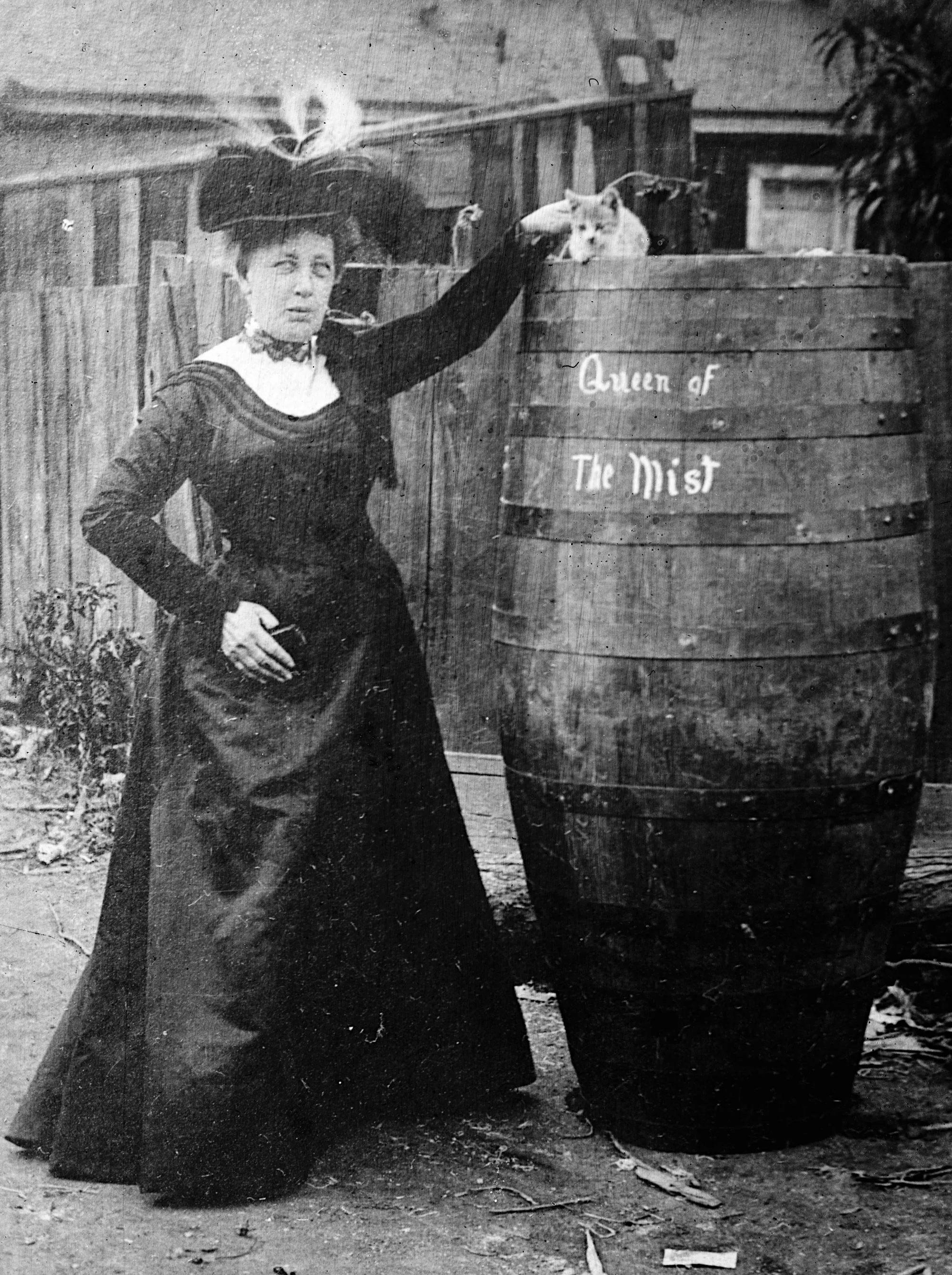 A woman in a dark dress and feathered hat stands beside a tall barrel marked Queen of the Mist, with a cat sitting on top.