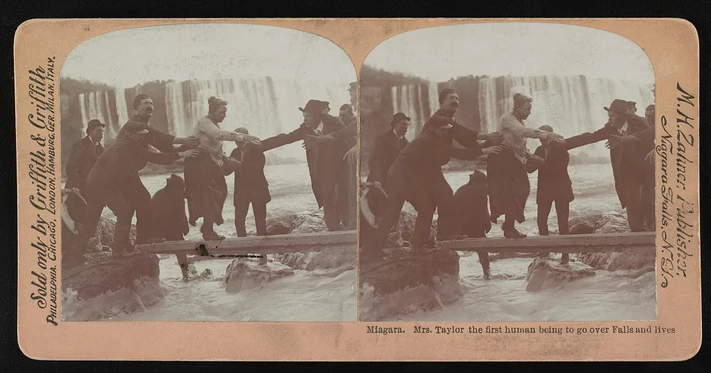 A sepia stereograph presents paired views of a woman stepping along a plank with men reaching toward her and a waterfall behind.