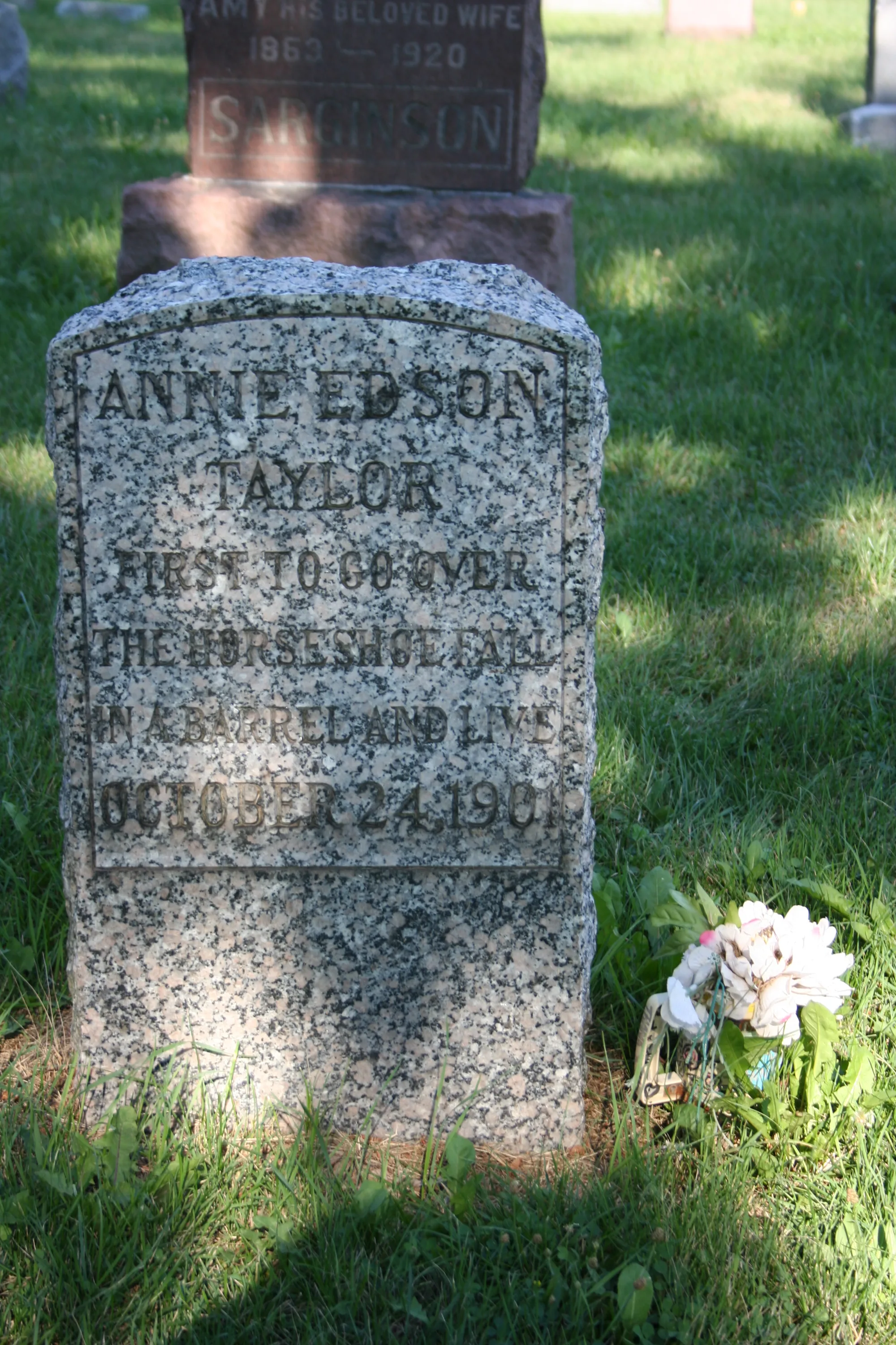 A grey grave marker inscribed Annie Edson Taylor stands in grass, with flowers beside it and another headstone behind.
