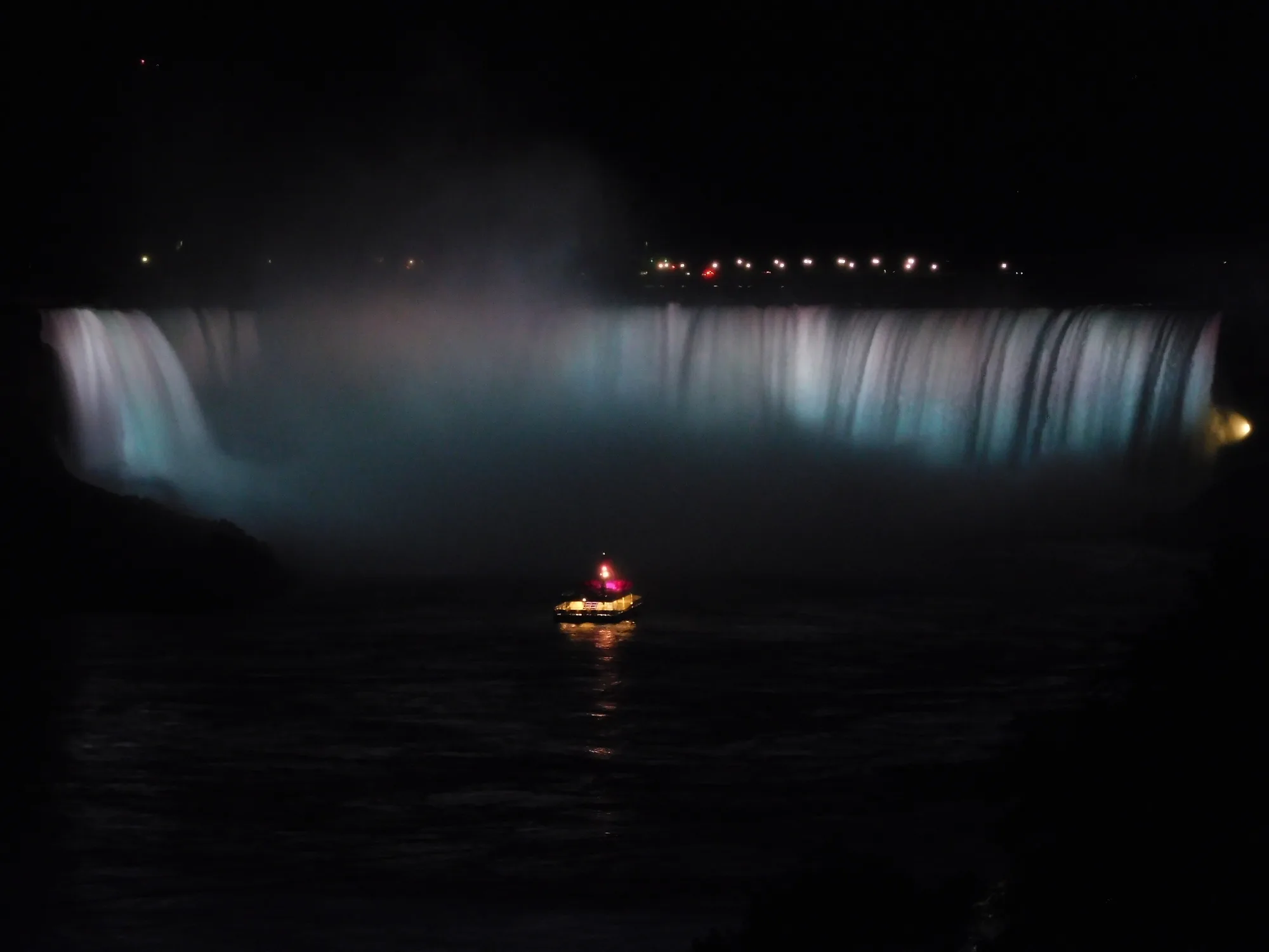 An illuminated curved waterfall shines pale blue and white at night, with a small lit boat in the dark water below.