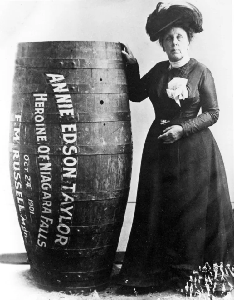 A woman in a dark dress and hat stands beside a barrel painted with her name, Heroine of Niagara Falls and the date October 24, 1901.
