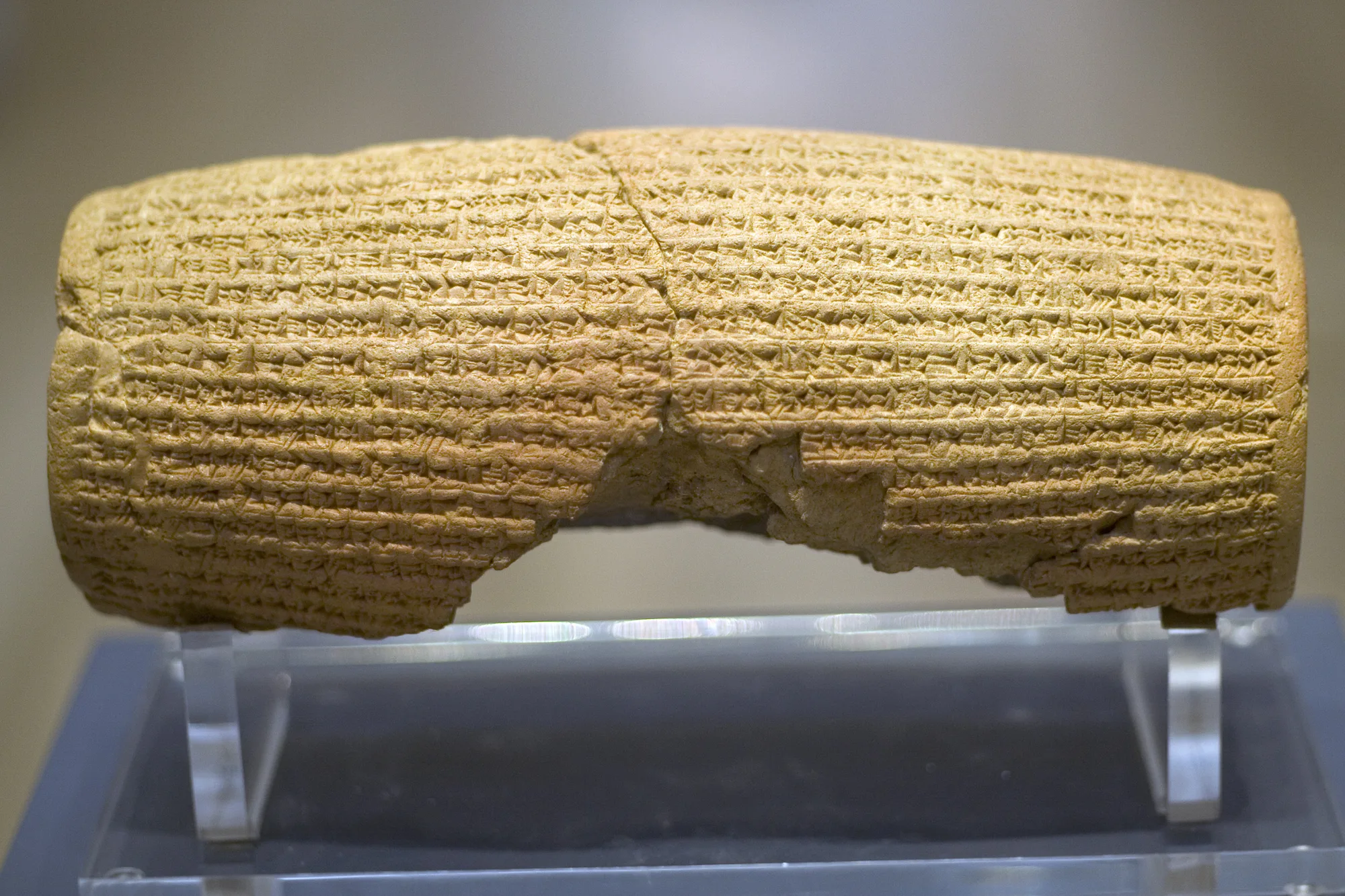 The reverse of a clay cylinder is covered in horizontal rows of writing, above a clear support and a chipped lower edge.
