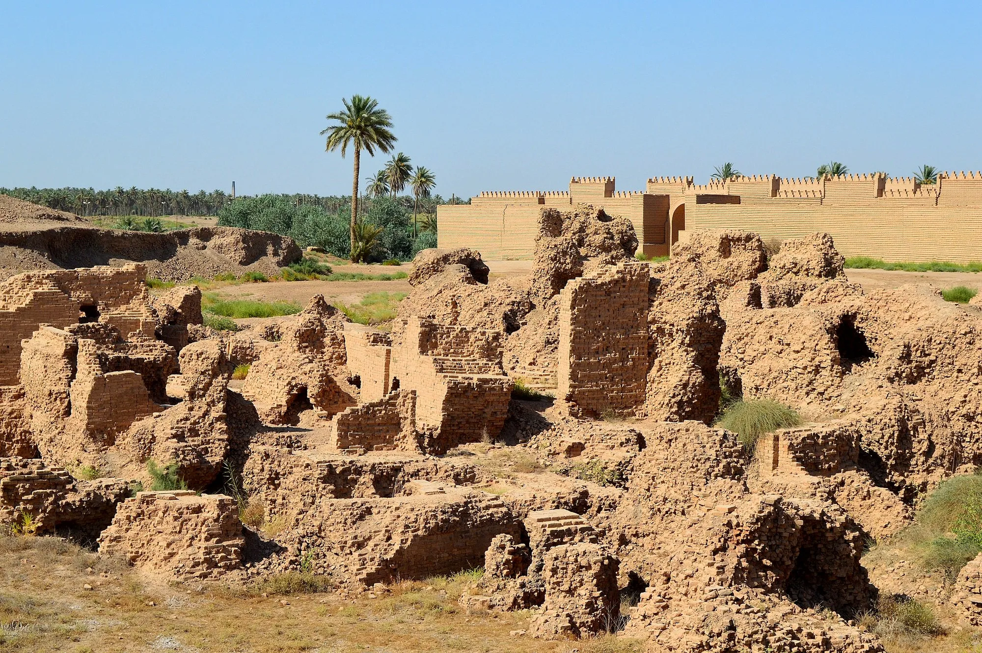 Low, eroded earthen walls and excavated spaces stand in front of a palm tree and a taller pale brick wall under a blue sky.