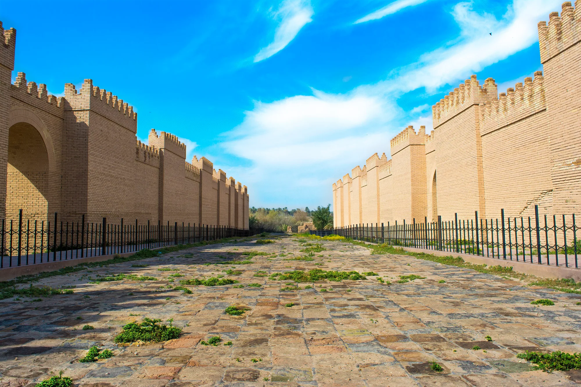 A paved path with patches of grass runs between tall pale brick walls with crenellated tops and black metal fencing.