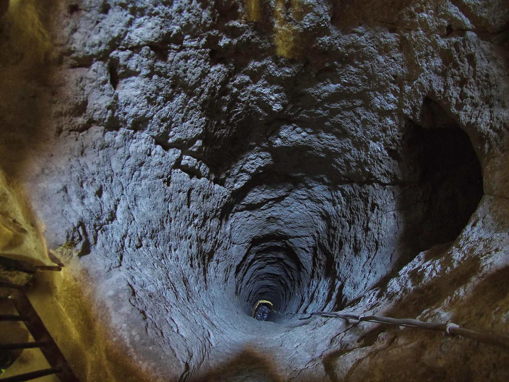 A view down a deep, rough-walled rock shaft, with a coil of cable on the near ledge and a narrow opening below.