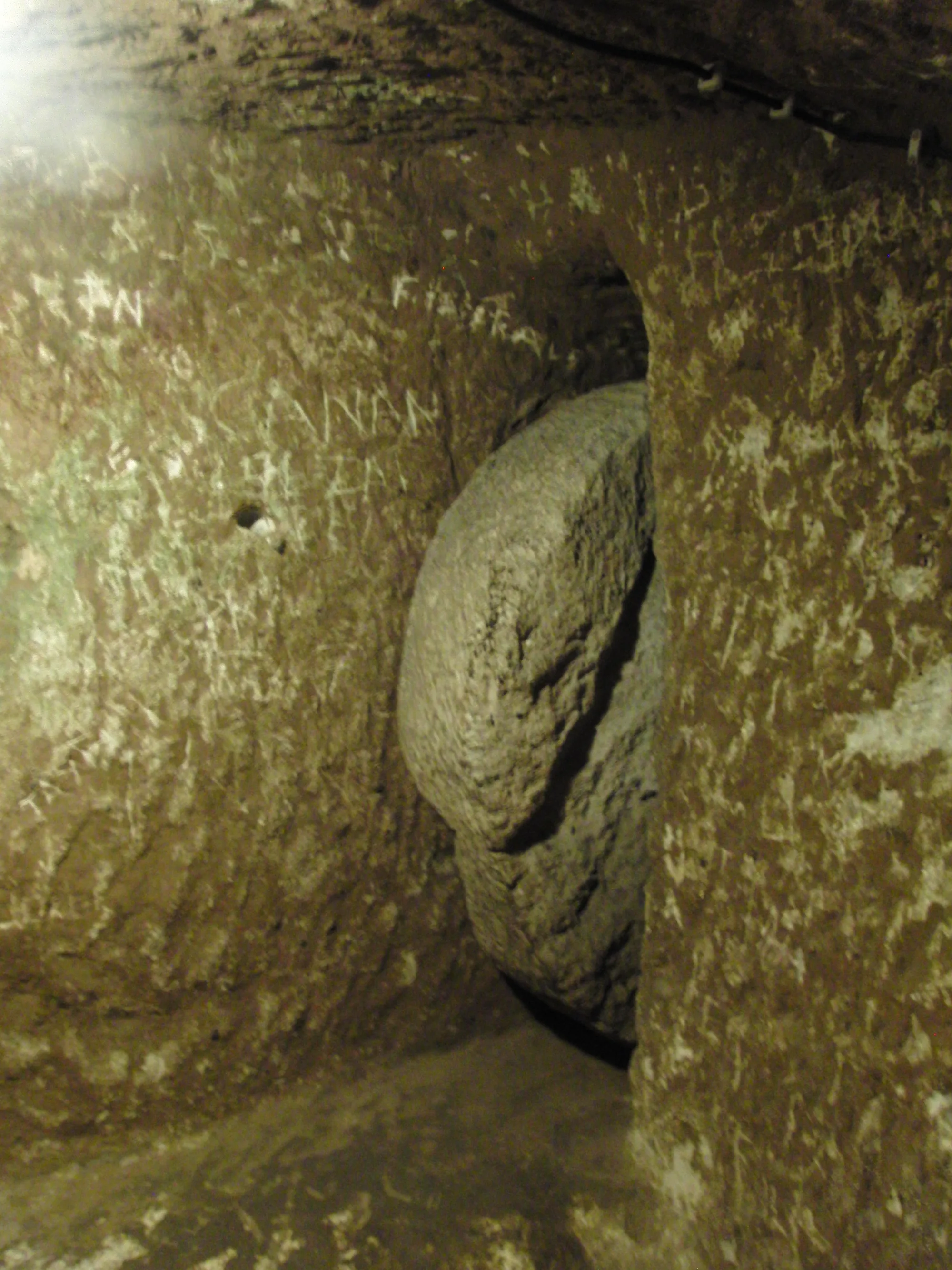 A heavy disc of stone, like a millstone standing on edge, set into a recess beside a narrow rock passage whose walls are scratched with graffiti.