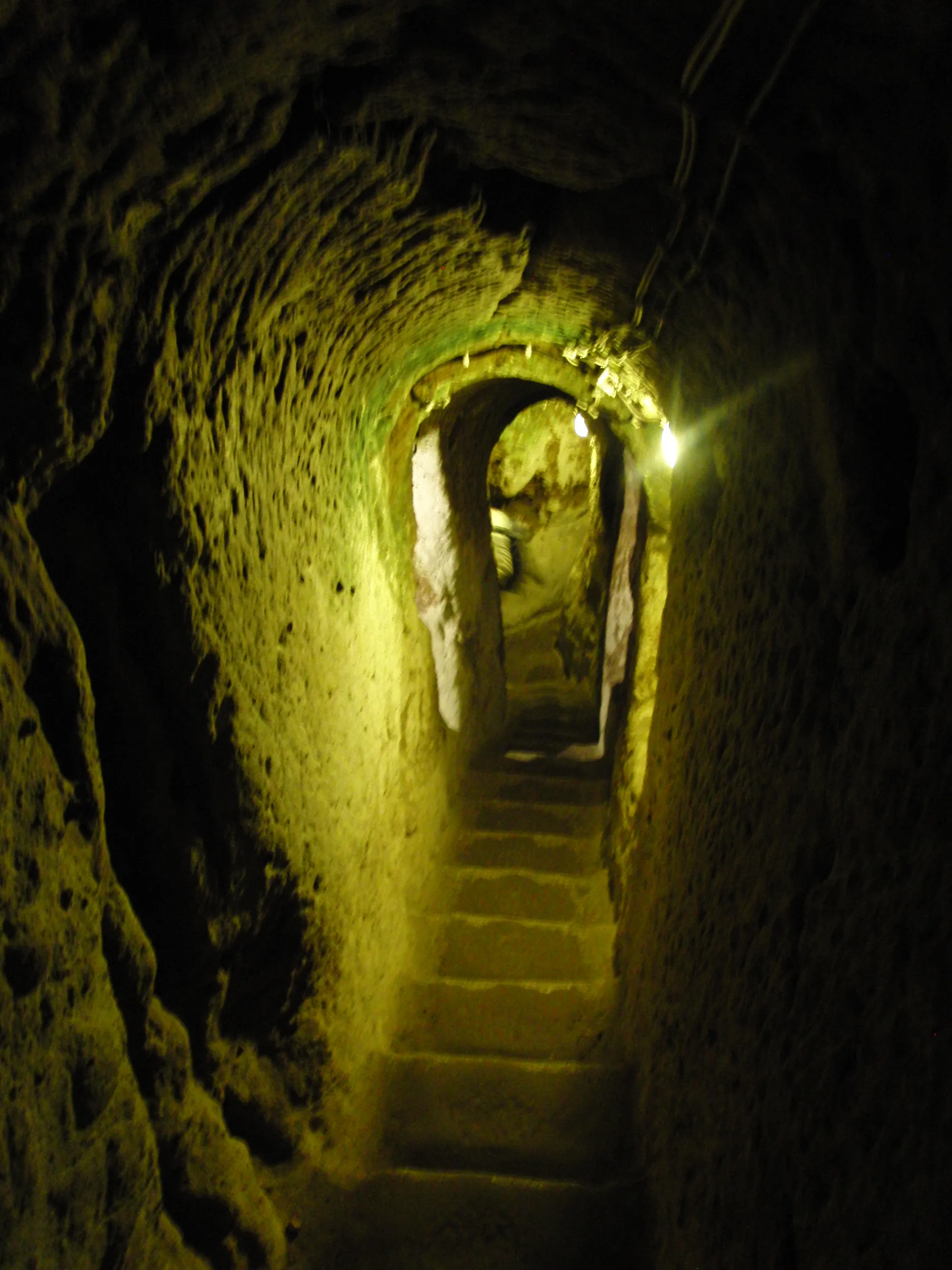 A narrow staircase carved from rock descends through a low arched tunnel lit by electric lamps.