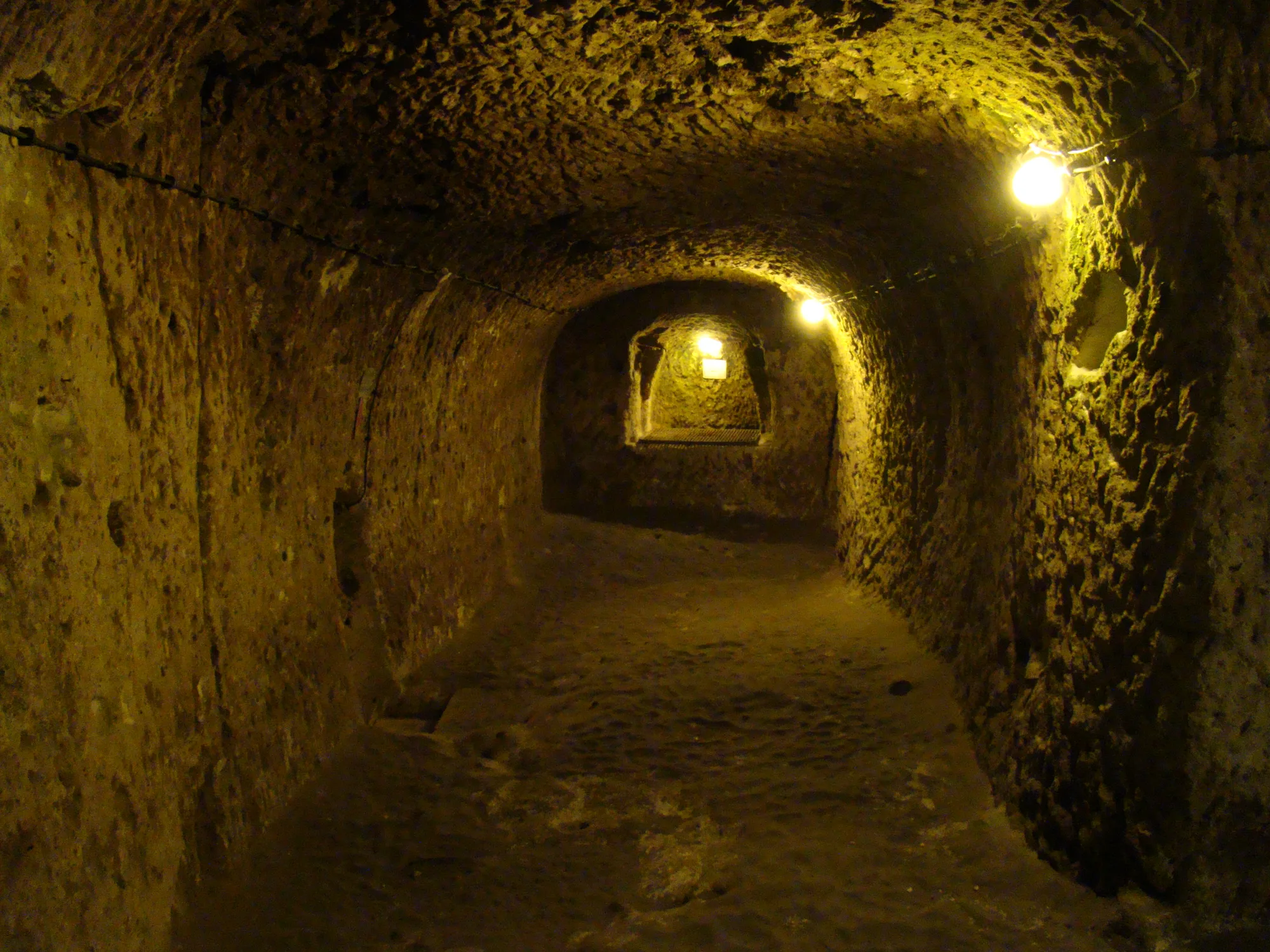 A wide, low corridor hollowed out of yellowish rock, lit by a few bulbs, opening into a further chamber at the far end.
