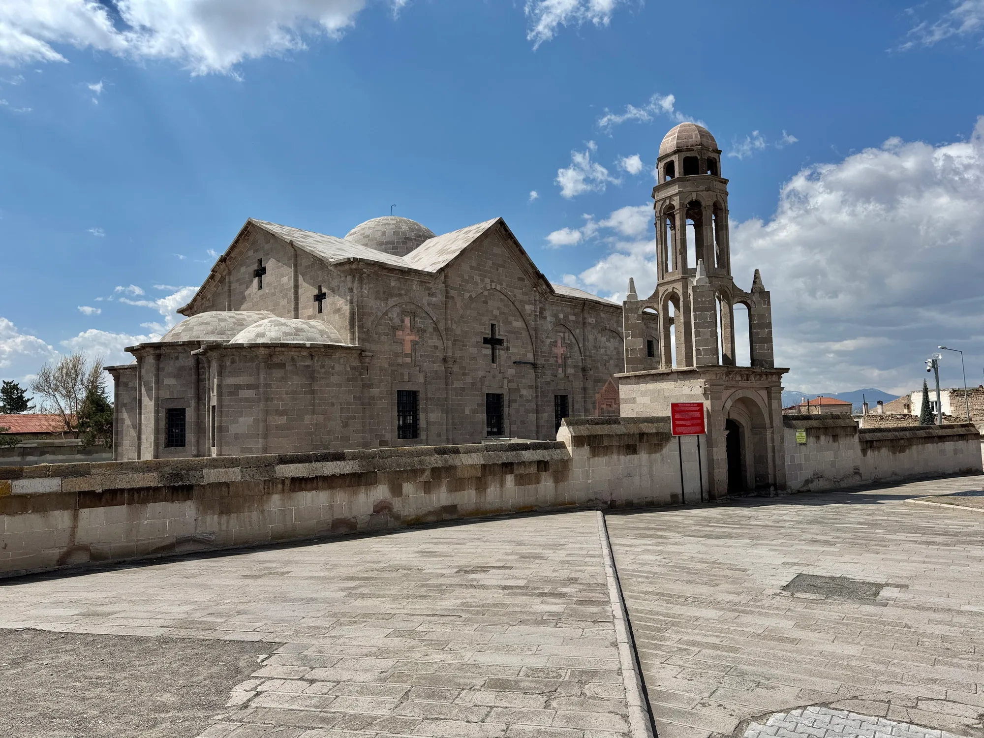 A stone church with pitched roofs and a tall arched bell tower topped by a small dome, behind a low wall beside a paved square.