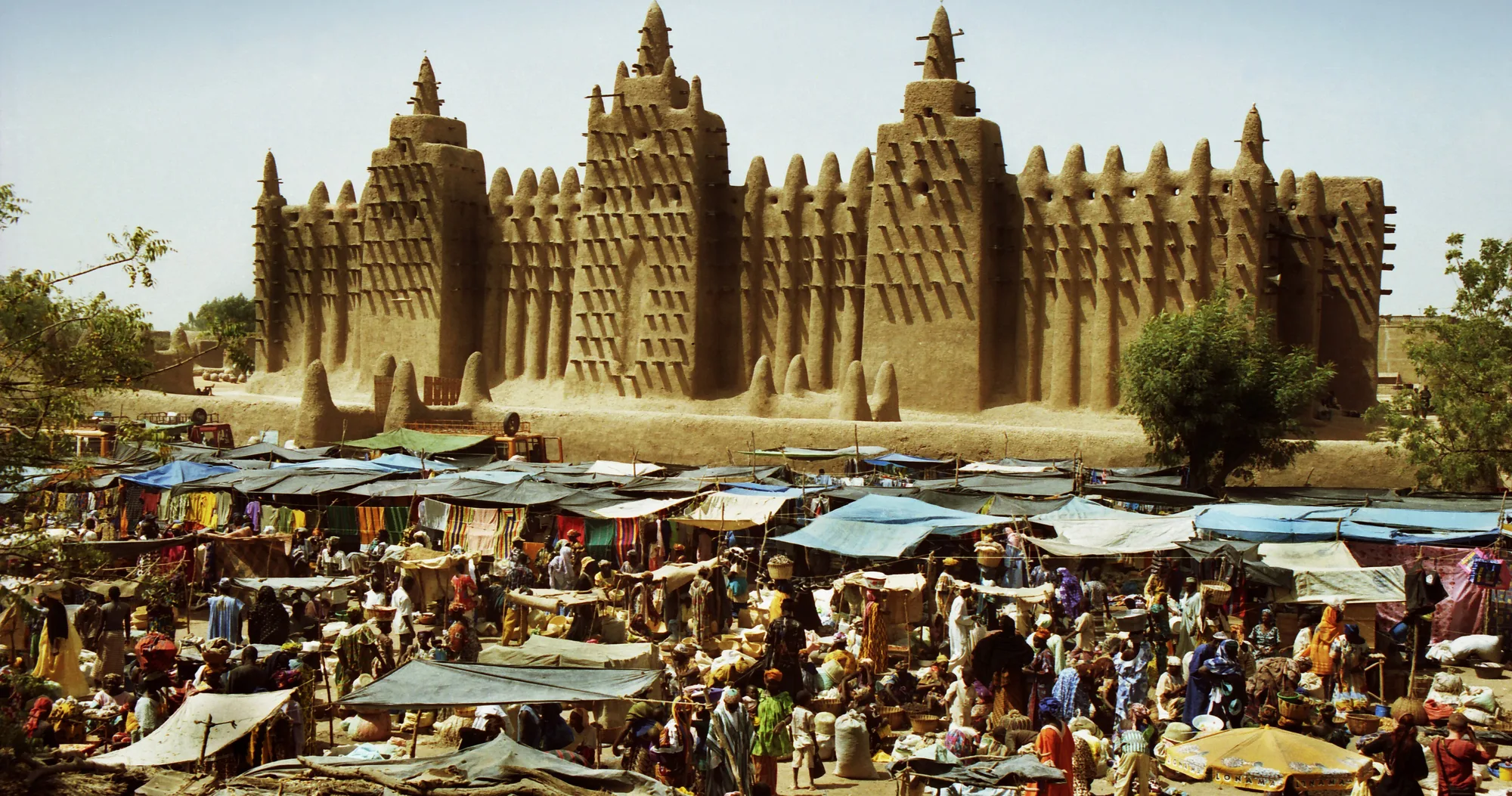 The long earthen wall of the Great Mosque of Djenné, with three tall towers bristling with wooden beams, rising above a crowded market of awnings and stalls.