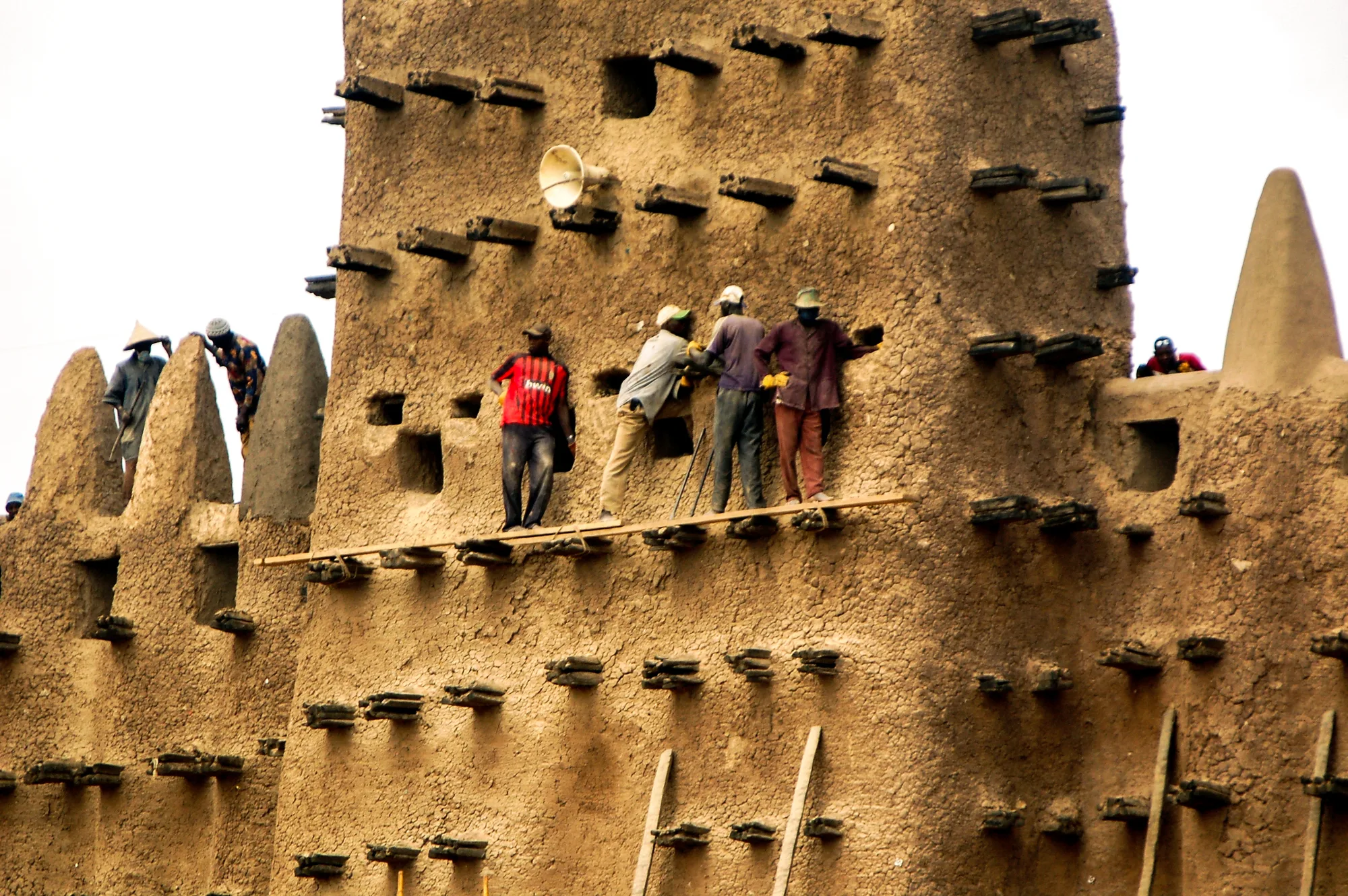 Men stand on a plank laid across the palm-wood beams that stick out of a mud tower, smoothing fresh plaster onto the wall high above the ground.