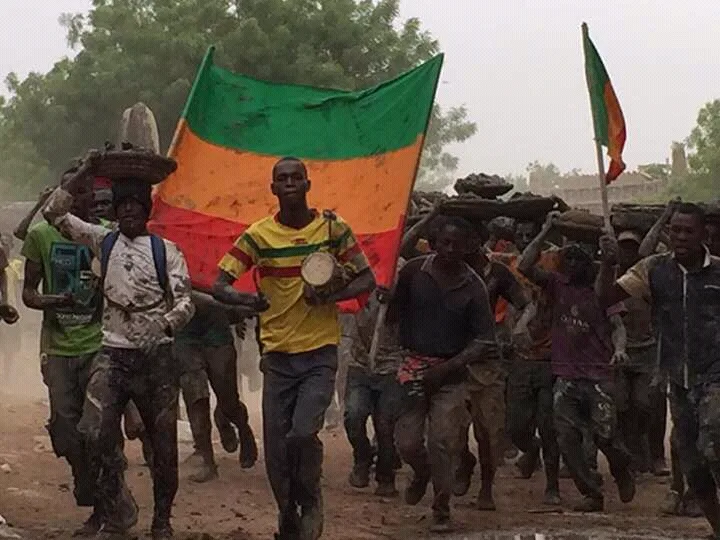 Men in mud-stained clothes carry loaded containers on their heads beside a drummer, with a large green, yellow and red cloth behind them.