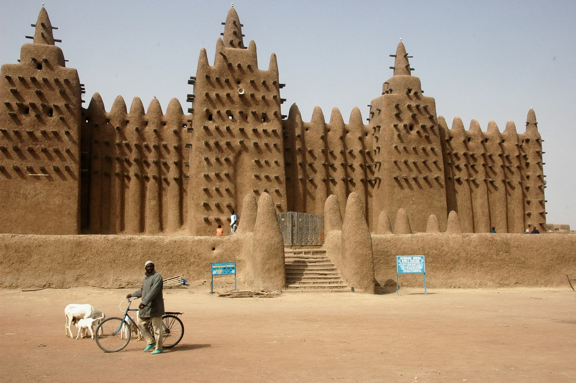 A man with a bicycle and two goats stands on a sandy square before the mosque's entrance stairway and its rows of rounded mud buttresses.