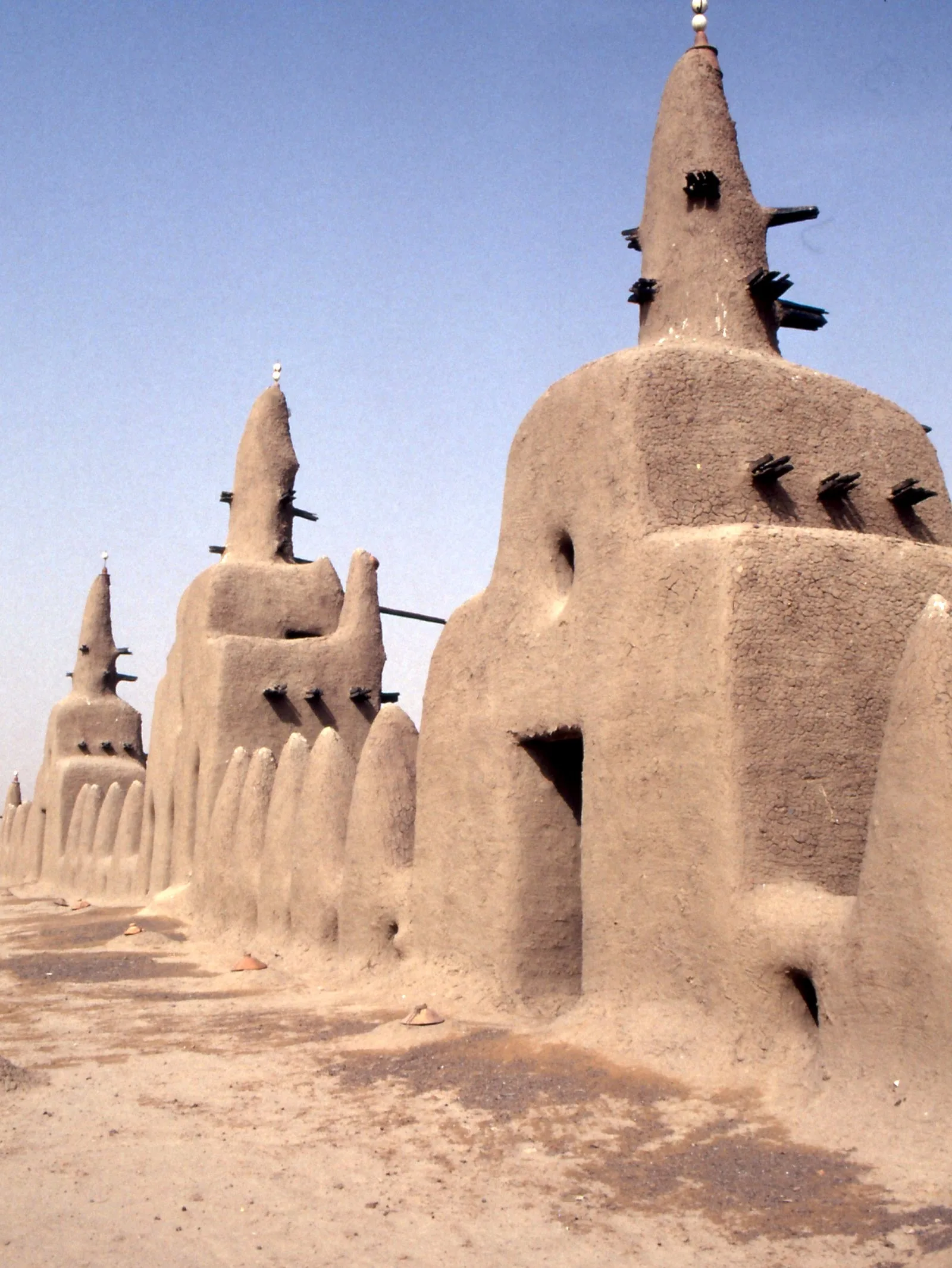 A view along the mosque roof toward tapering earthen towers, with wooden beams projecting from their walls and small terracotta covers along the flat roof.