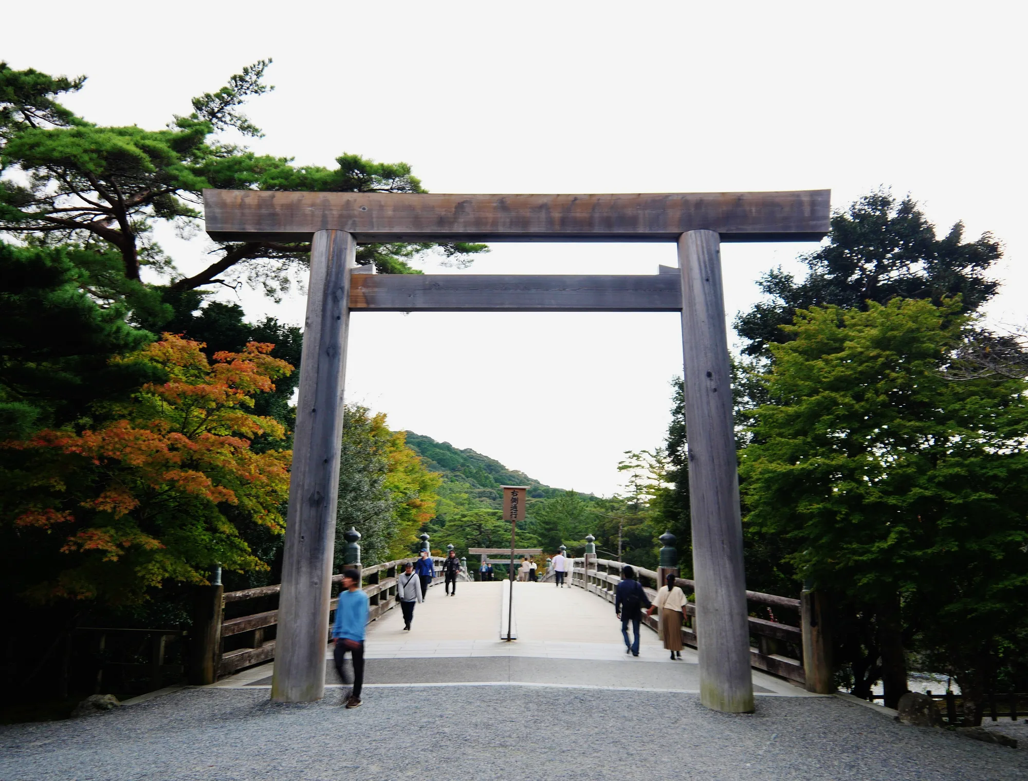 A tall plain wooden torii gate frames the entrance to a long wooden footbridge, with visitors walking across toward forested hills.