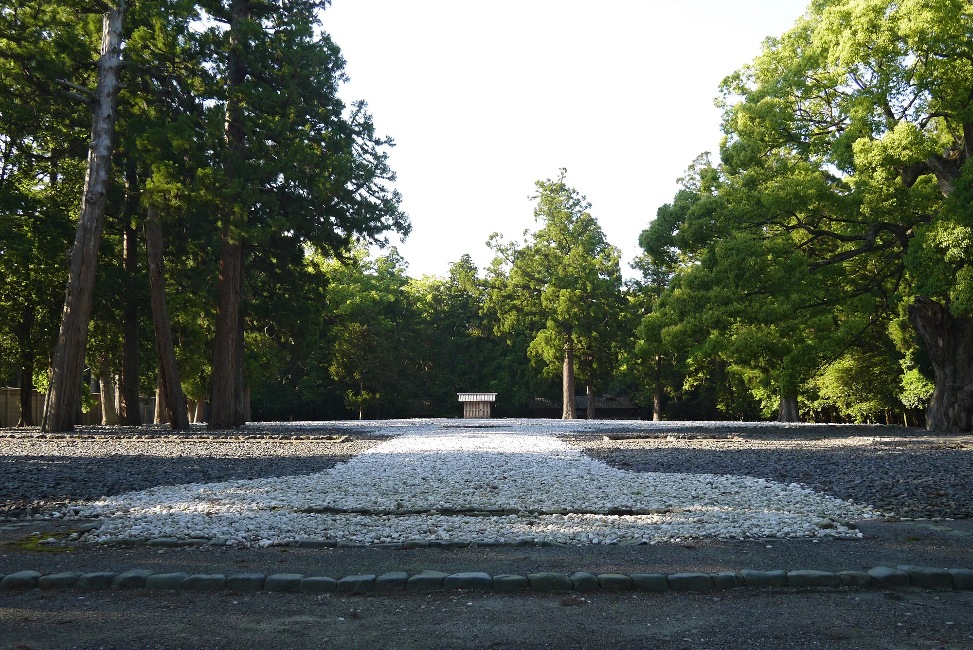 A wide rectangle of white and grey pebbles in a clearing surrounded by tall trees, empty except for a small wooden hut in the middle.