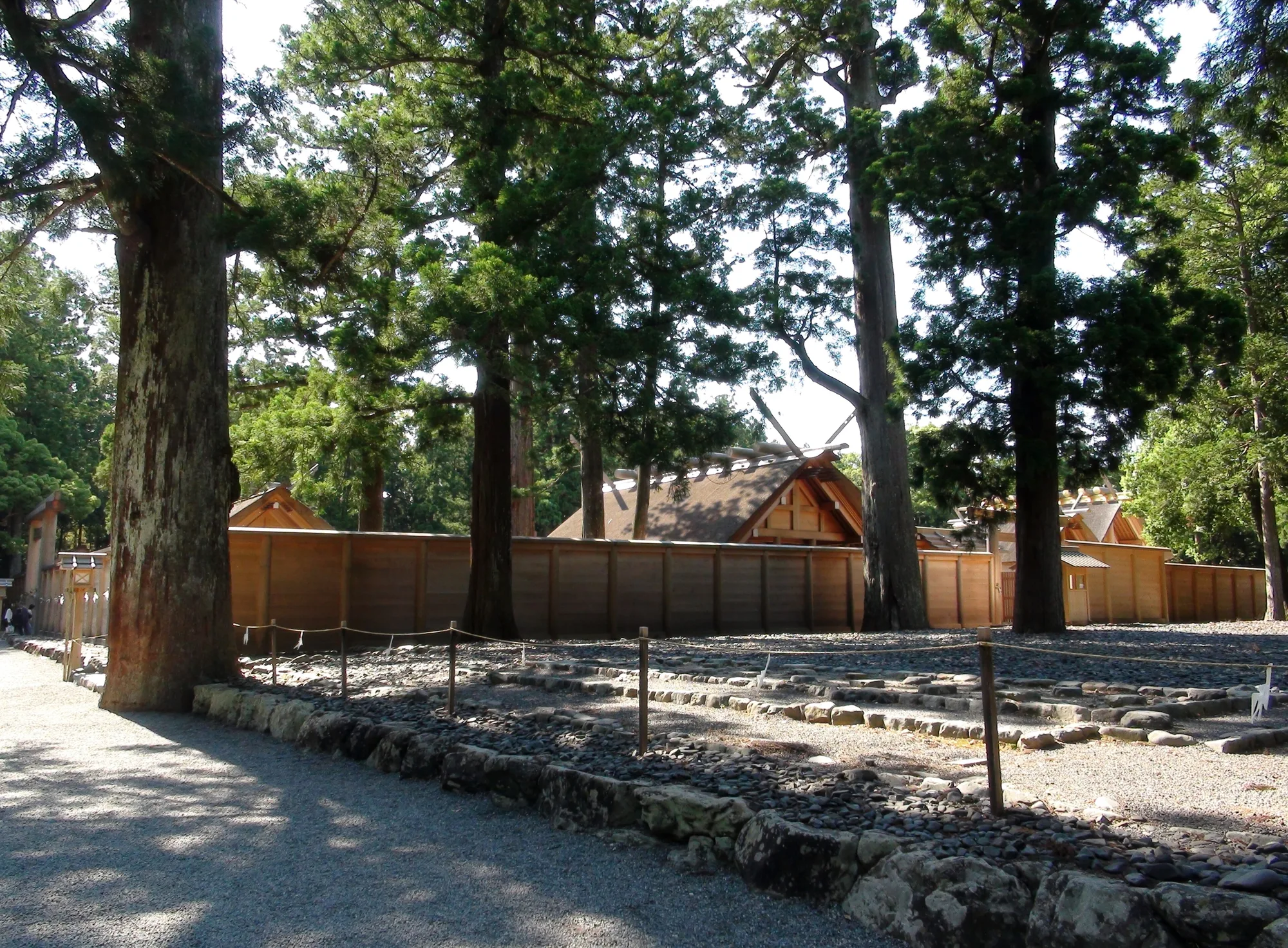 Thatched roofs rising behind tall plain wooden fences among old cedar trees, with a roped-off field of pebbles in the foreground.