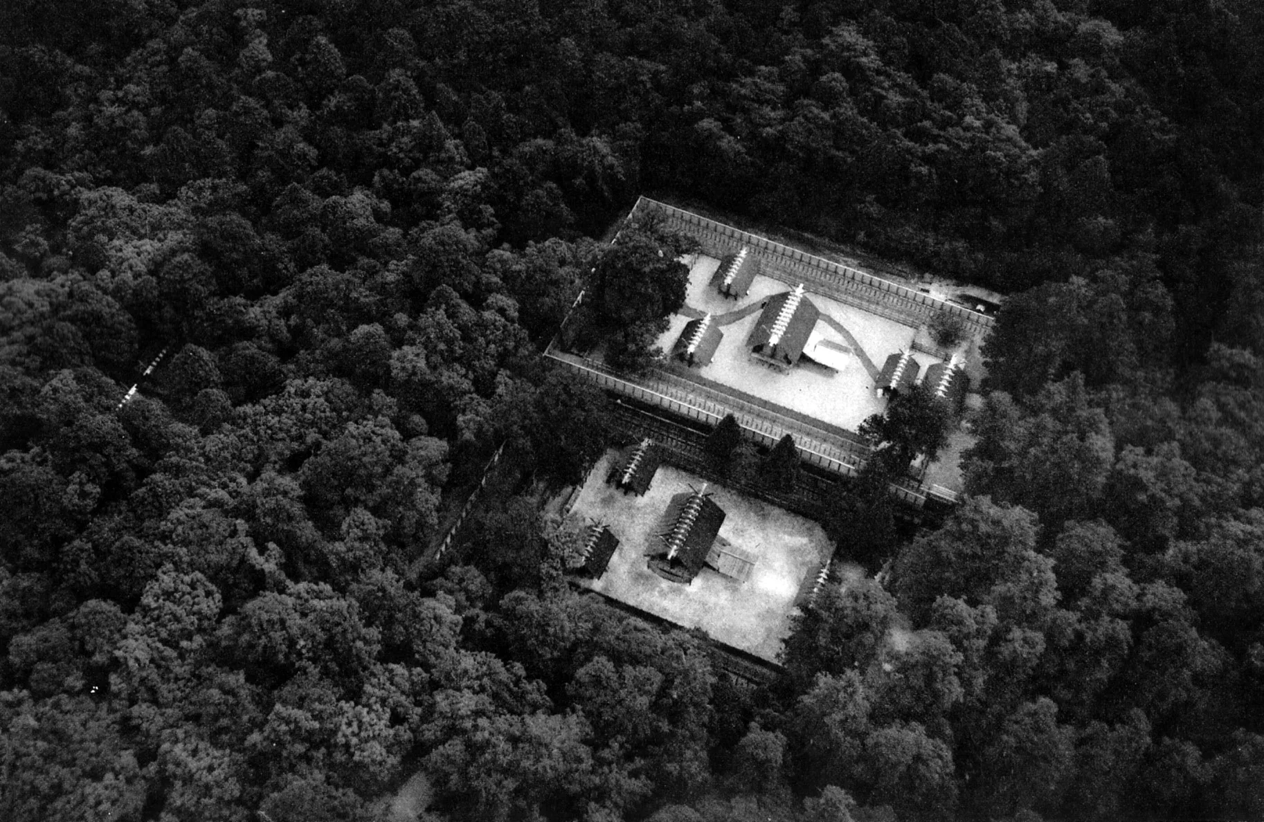 Black-and-white aerial photograph of two rectangular fenced shrine compounds side by side in a dense forest.