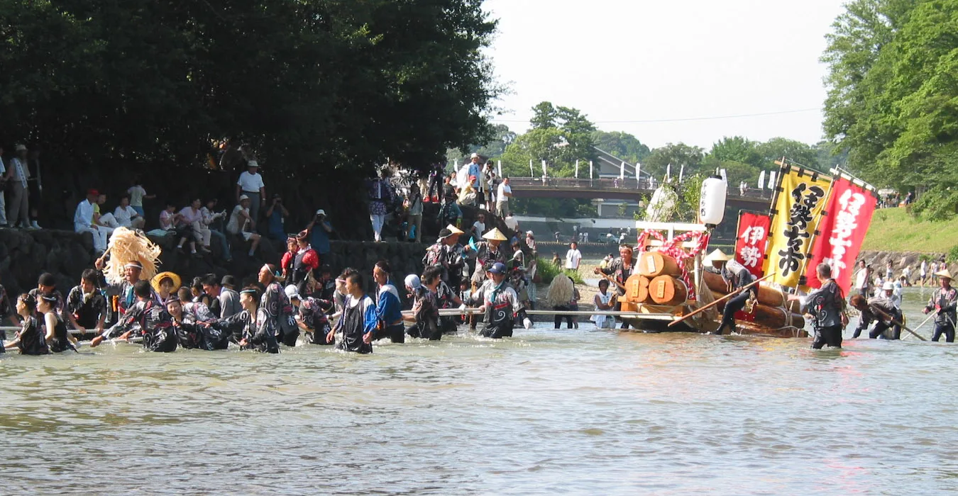 A crowd of people waist-deep in a shallow river hauling a sled stacked with timber and decorated with banners, while spectators watch from the bank.