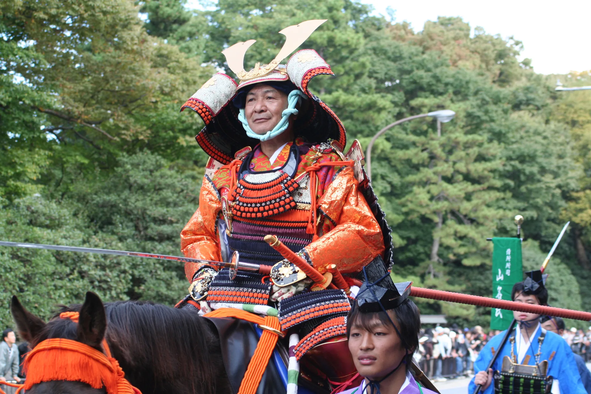 A rider in bright orange armour and a crested helmet sits on a horse beside an attendant, with spectators lining the street.