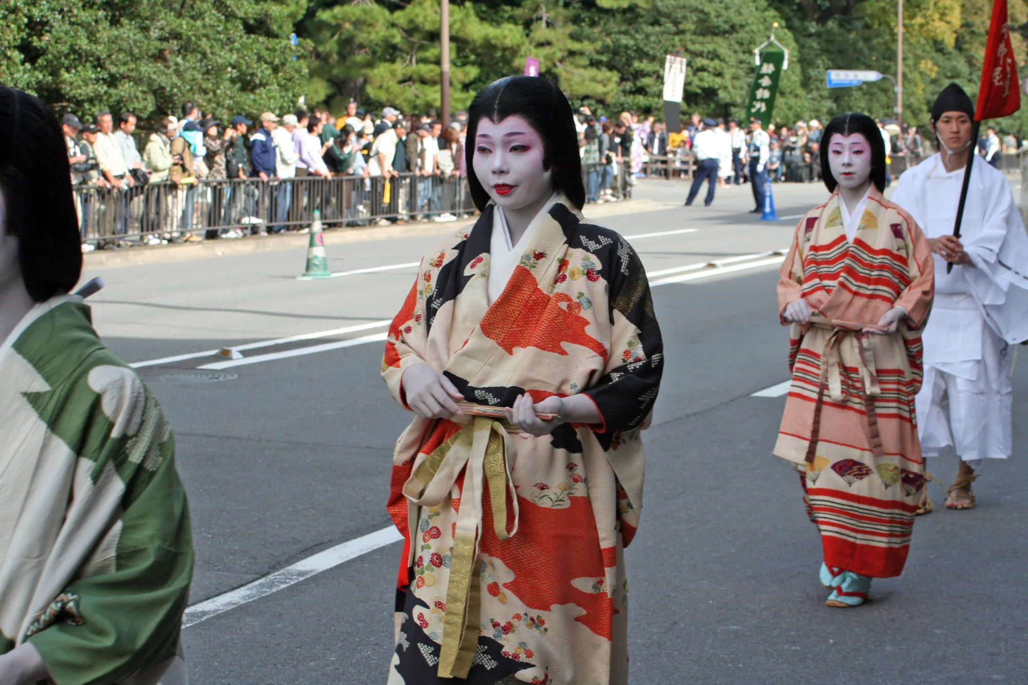 Two participants in patterned robes and white face makeup walk along a street beside spectators behind railings.