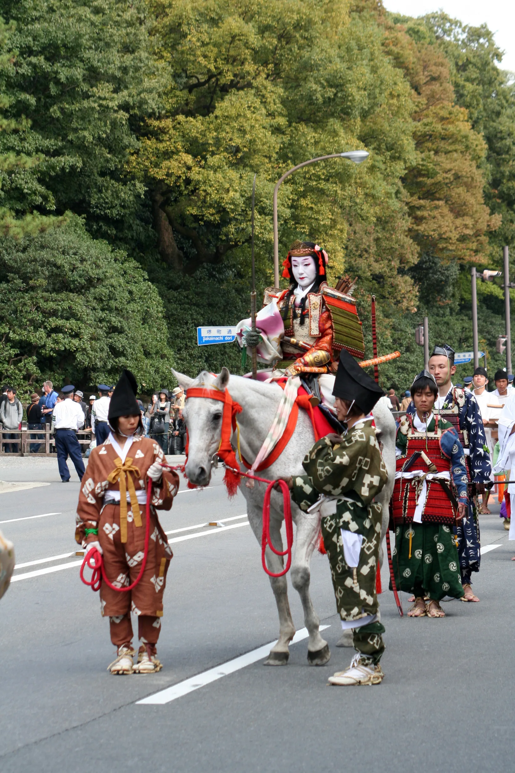An armoured participant rides a white horse led by attendants in a street procession.