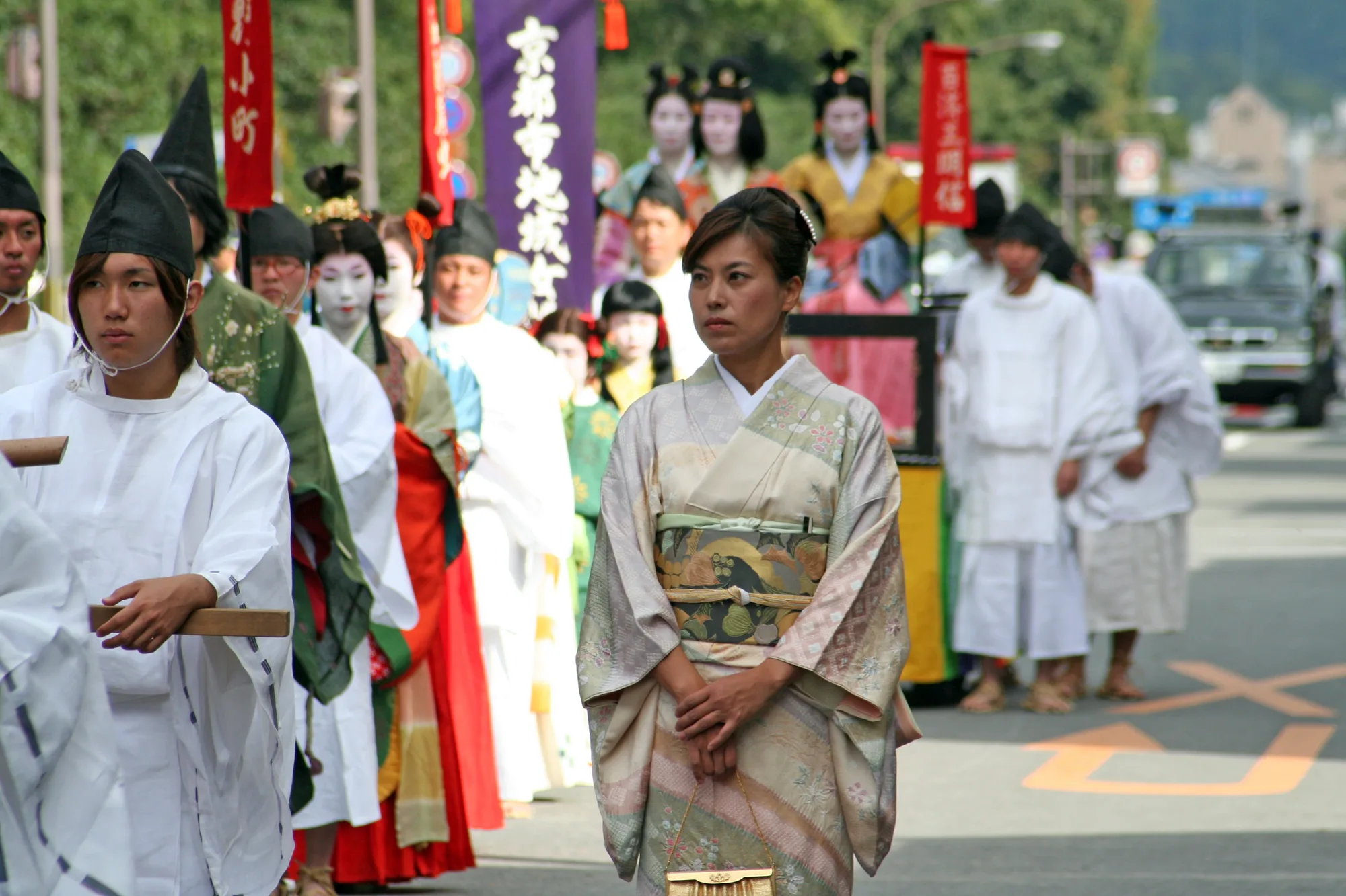 A participant in a pale patterned robe stands beside white-robed attendants, with brightly dressed figures and banners behind.