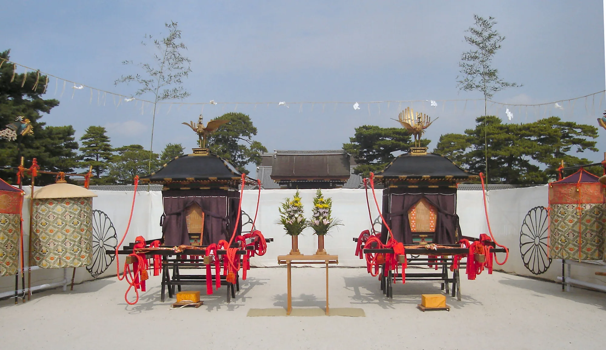 Two black-roofed carriages with gold bird-shaped finials and red carrying poles stand before white screens, with a small flower stand between them.