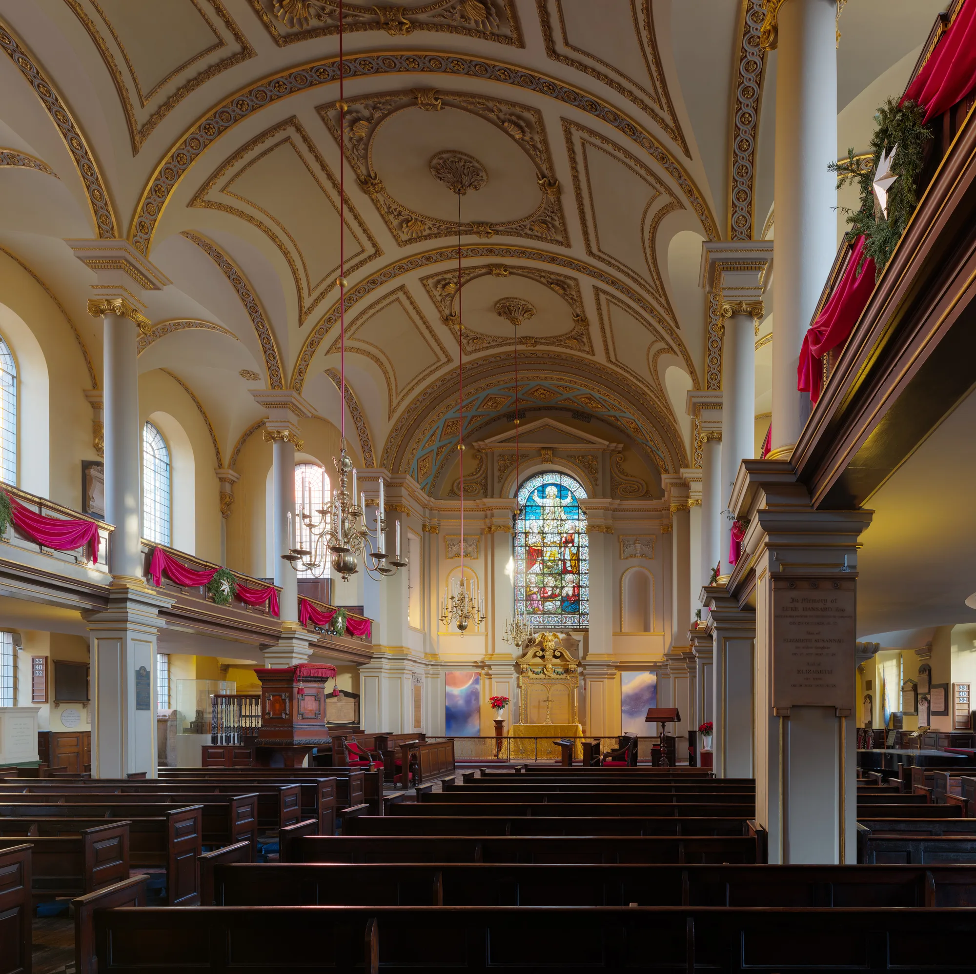 A church interior with rows of dark wooden pews, pale columns and galleries on both sides, looking toward a tall east window.
