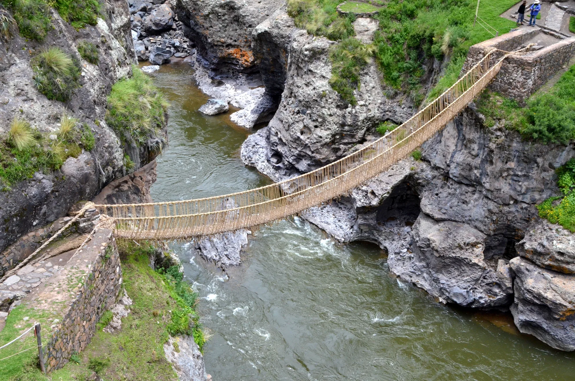 A sagging bridge of woven straw-coloured grass rope hangs between stone abutments across a rocky gorge, with a fast green river below.