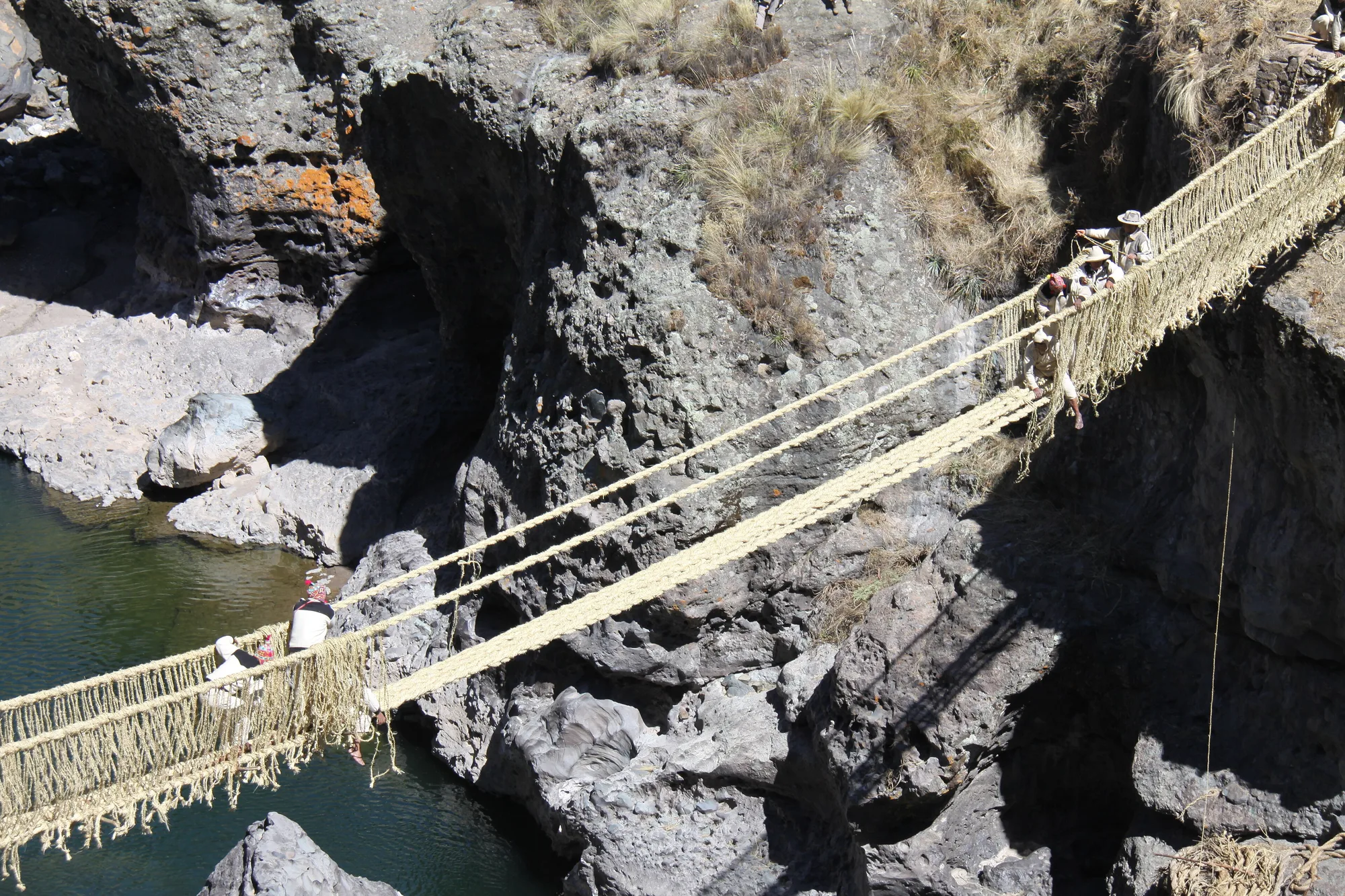 Seen from above, men in hats stand on the grass-rope bridge high over the river as they work on its side netting, while another group works near the far end.