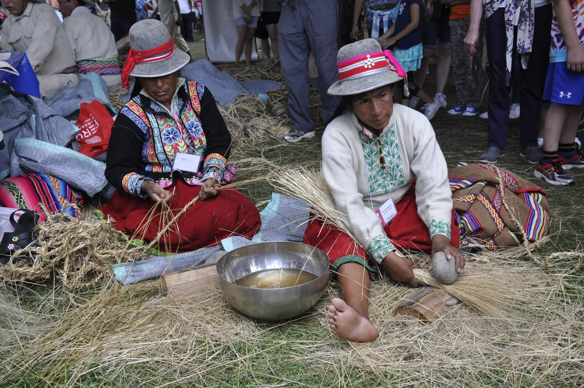 Two Quechua women in embroidered jackets and felt hats sit on the grass among bundles of straw; one twists fibres while the other pounds a bundle with a stone beside a metal bowl of water.