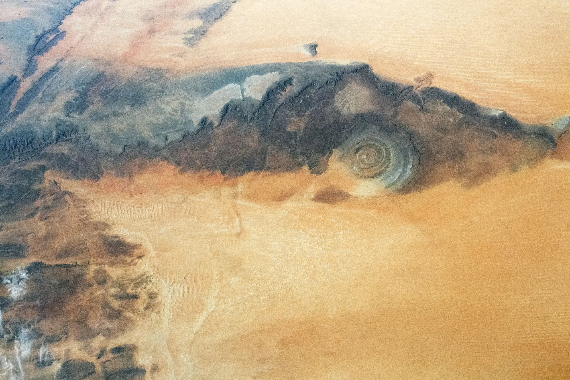 A circular set of pale and dark rock rings lies within a dark plateau bordered by broad golden dune fields.