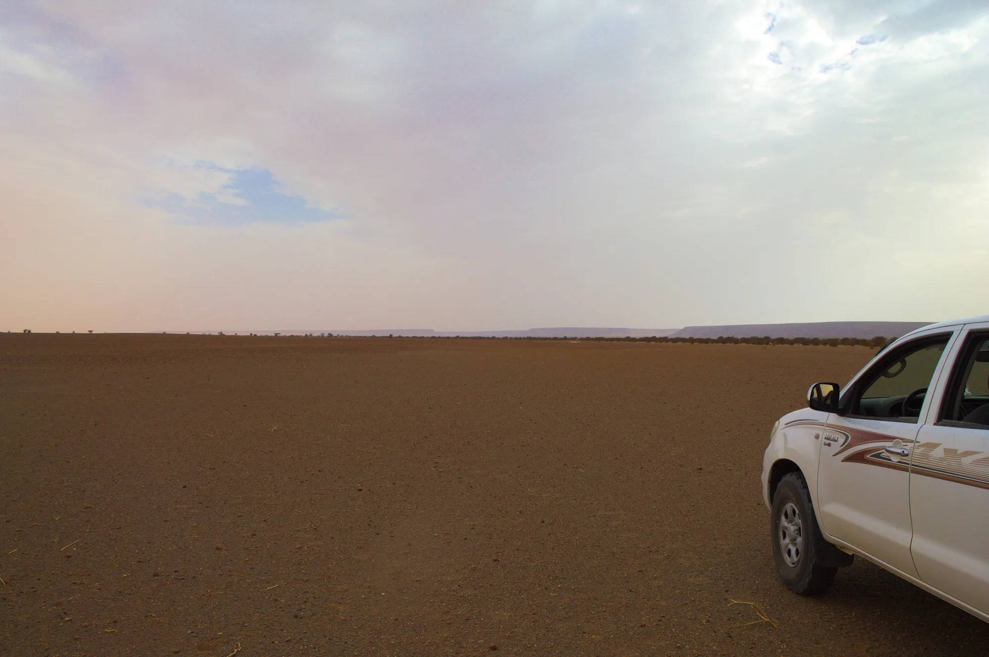 A flat brown gravel plain extends to low distant ridges beneath a cloudy sky, with part of a white vehicle at the right edge.