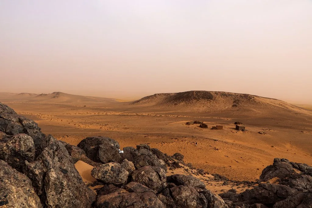 Jagged rocks in the foreground overlook a sandy basin, low dark-topped ridges and small structures under a hazy sky.