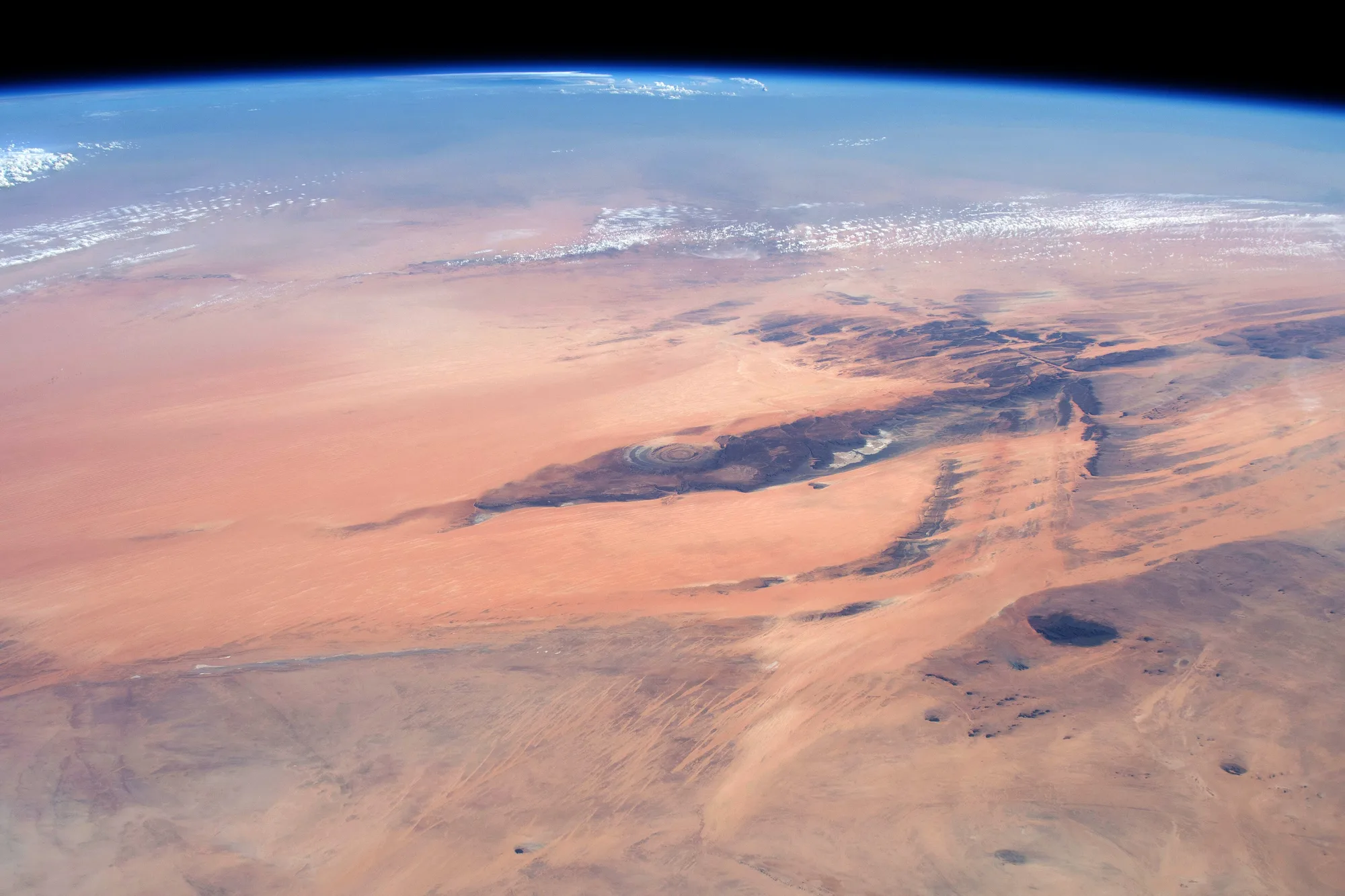 A broad oblique view shows dark ridges and a small ringed formation amid pale desert, below a blue atmospheric edge and scattered clouds.