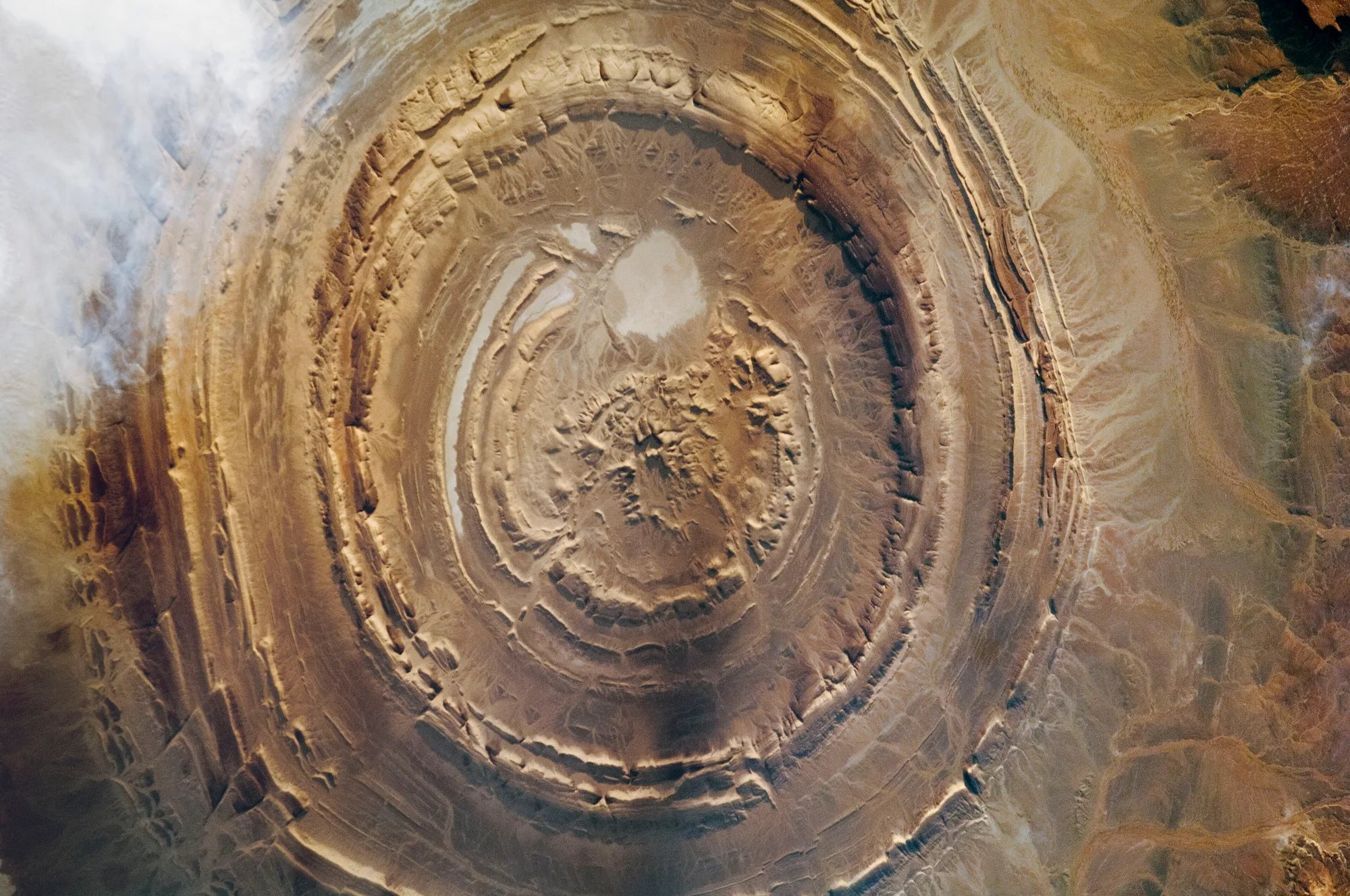 Concentric pale ridges and darker intervening bands surround a rough central area, with cloud obscuring part of the left side.