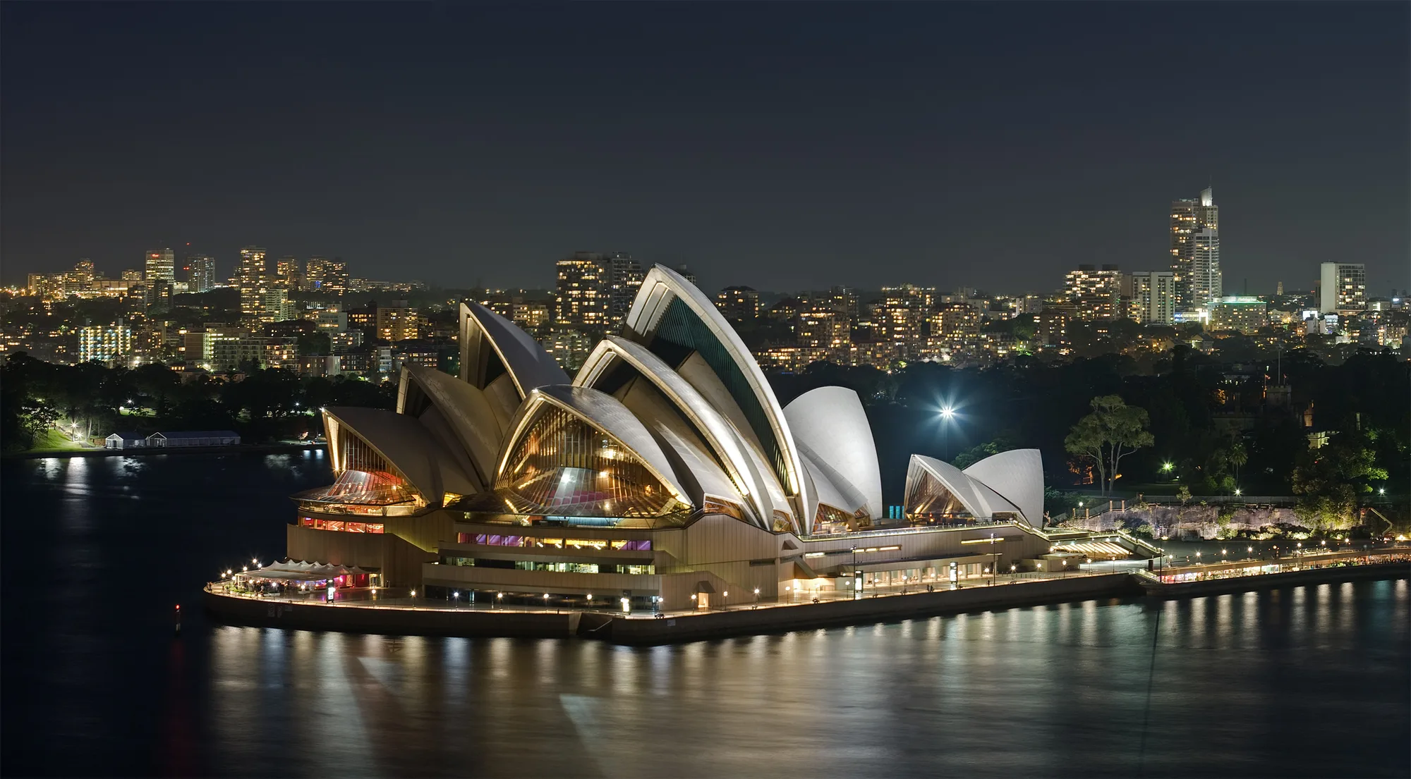 The Sydney Opera House is lit at night on a waterfront platform, with its roofs reflected in dark harbour water.