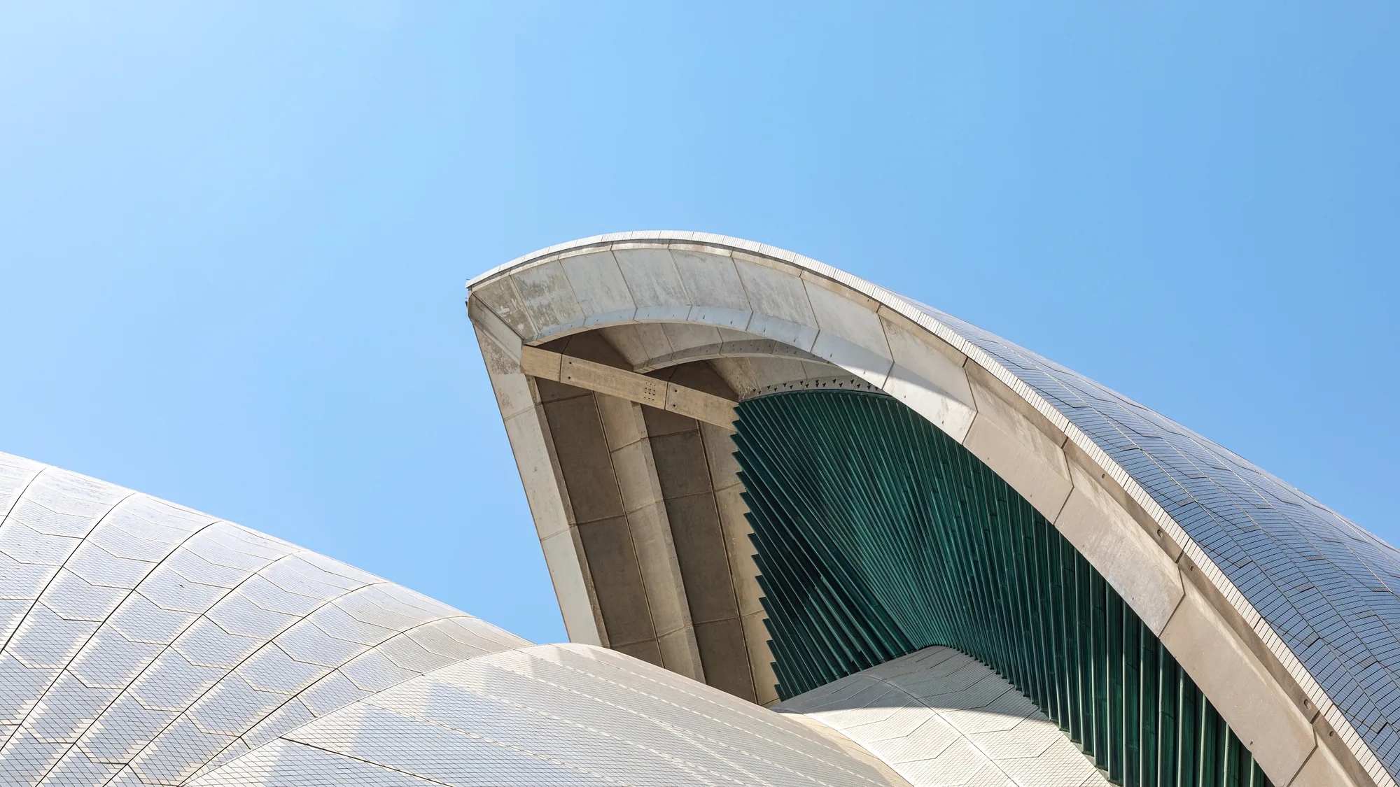 A close view of curved tiled roof shells and green glass beneath a blue sky at the Sydney Opera House.