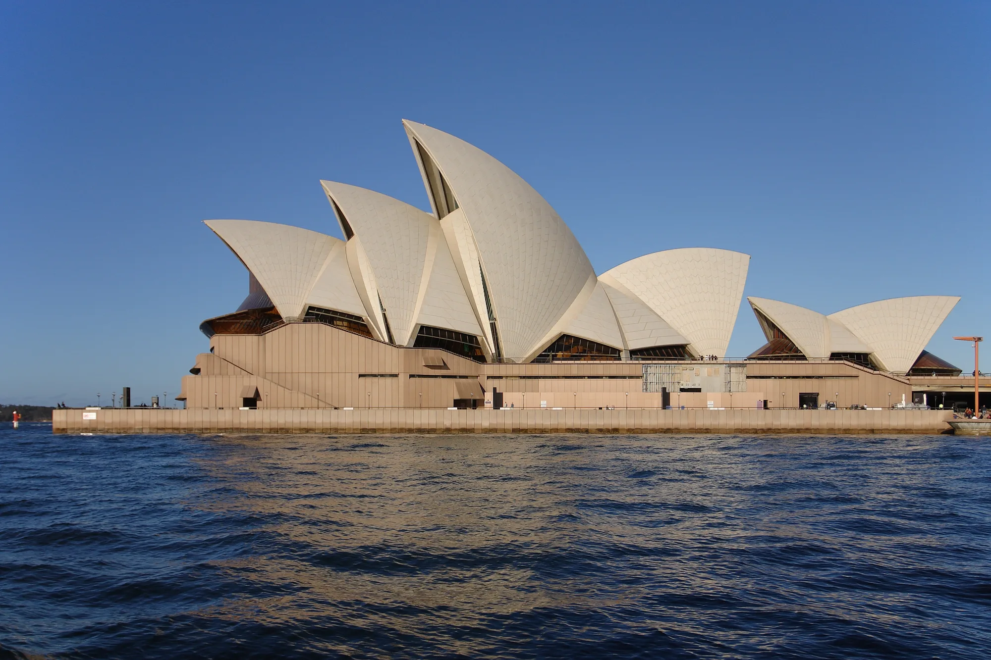 The Sydney Opera House’s white roof shells and pale waterfront base are seen across blue harbour water in daylight.