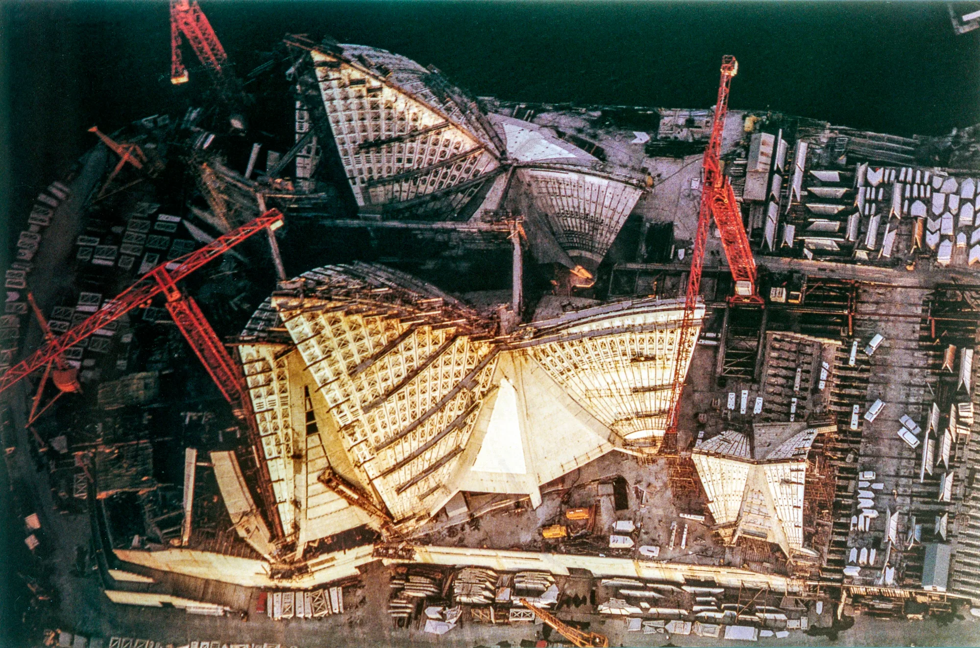 An aerial view of the Sydney Opera House under construction, with partly built ribbed roof shells, red tower cranes and stacked precast segments across the site.