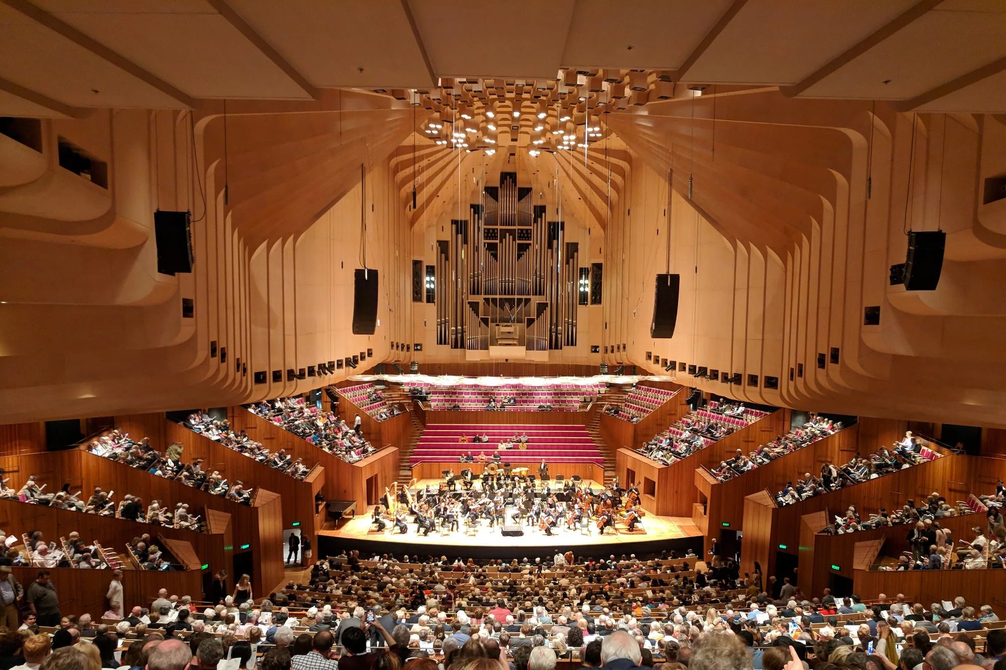 An audience fills tiered seating around a stage with musicians and instruments, beneath pale ribbed walls and a pipe organ.